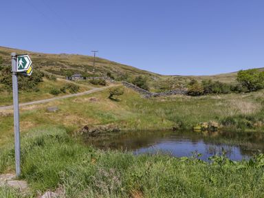A grassy landscape with a pond a walking path and a walking direction signpost at Llethr - Shepherds Hut in Llanbedr