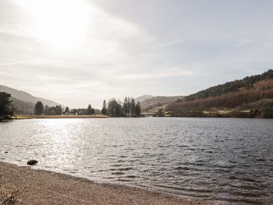 A lake with a pebble beach surrounded by trees and hills at Owl Lodge in South Laggan near Invergarry