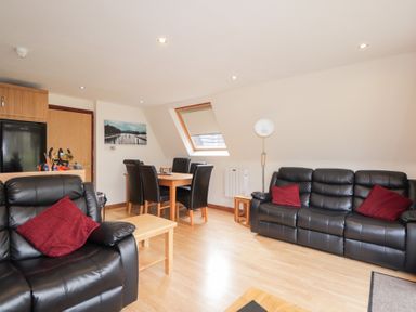A living room with black leather sofas and red cushions a wooden dining table with black chairs and a wooden floor at Owl Lodge in South Laggan near Invergarry