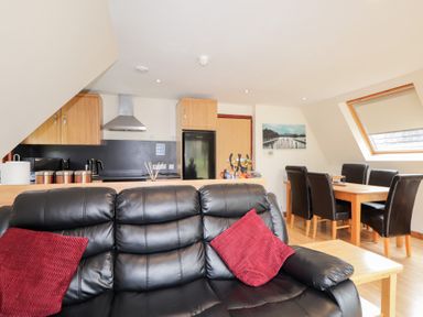A living room with a black leather sofa and red cushions next to a wooden dining table with six dark chairs and a kitchen with wooden cabinets and black appliances at Owl Lodge in South Laggan near Invergarry