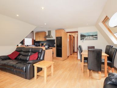 A living area with a black leather sofa and red cushions next to a wooden side table a dining table with four black leather chairs and an open kitchen with wooden cabinets at Owl Lodge South Laggan near Invergarry
