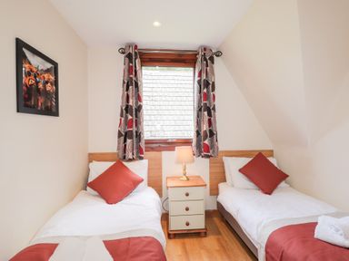 A bedroom with two single beds separated by a nightstand with a lamp and a window with curtains at Owl Lodge in South Laggan near Invergarry