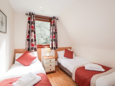 A bedroom with two single beds a nightstand with a lamp and a window with patterned curtains at Owl Lodge South Laggan near Invergarry