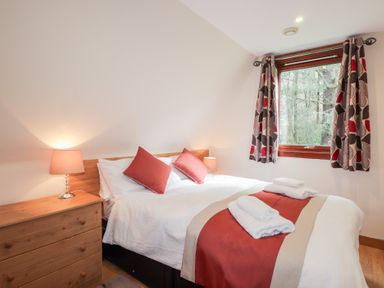 A bedroom with a double bed bedside lamps a wooden chest of drawers and a window with patterned curtains at Owl Lodge in South Laggan near Invergarry