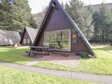 Two brown wooden A frame cabins with picnic tables outside on grass surrounded by trees at Owl Lodge South Laggan near Invergarry
