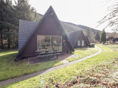 A row of wooden a frame cabins with picnic tables on brick patios beside a curved path at Owl Lodge South Laggan near Invergarry