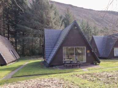 Triangular wooden cabins with dark roofs and large windows surrounded by grass and trees at Owl Lodge South Laggan near Invergarry