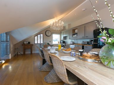 A kitchen with a long wooden dining table set with plates cups and food surrounded by wicker chairs a chandelier above and grey cabinets at 7 Four Seasons in Carbis Bay