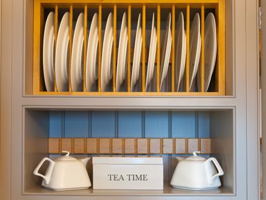 White plates on a wooden rack above two white teapots and a box labeled tea time on a shelf at 7 Four Seasons in Carbis Bay