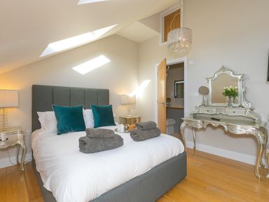 A bedroom with a bed with pillows and towels bedside tables lamps a vanity table with a mirror a chandelier and an open door to a bathroom at 7 Four Seasons in Carbis Bay