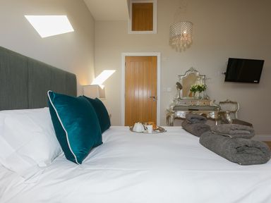 A bedroom with a made bed with teal pillows and towels on it a wooden door a decorative vanity table with mirror and flowers and a wall-mounted TV at 7 Four Seasons in Carbis Bay