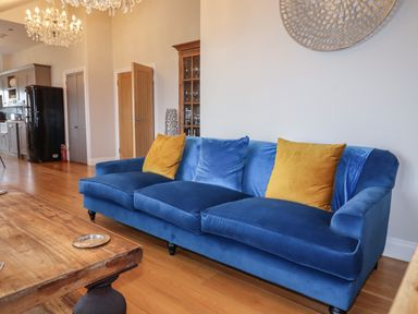 A blue sofa with yellow and blue cushions in a living room with a wooden table and kitchen area at 7 Four Seasons in Carbis Bay