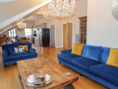 An open living room and kitchen area with blue sofas yellow cushions wooden coffee table and chandeliers at 7 Four Seasons in Carbis Bay
