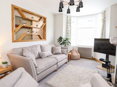 A living room with a grey sofa, a beige rug, a wall-mounted wooden shelf, a television on a stand, and a large window with patterned curtains at Dovestones in Keswick