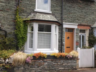 A stone house exterior with a bay window flowers and plants by the window and a wooden front door at Dovestones in Keswick