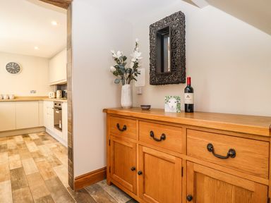 A wooden sideboard with a vase of flowers a small bowl a bottle of wine and a sheep picture next to a kitchen with white cabinets and wooden countertops at Dovestones in Keswick