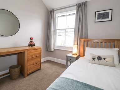 A bedroom with a wooden bed white bedding a bedside table with a lamp a wooden dresser with drawers a round mirror and a window with curtains at Dovestones Keswick