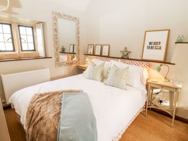 A bedroom with a bed pillows blanket bedside tables lamps and framed pictures near a window at The Small House Near Bourton-On-The-Water