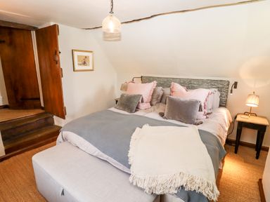 A bedroom with a bed with pink and gray pillows a light gray blanket and a small table with a lamp at The Small House Near Bourton-On-The-Water