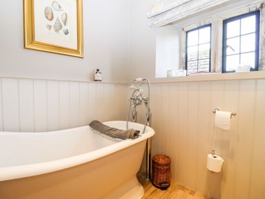 A bathroom with a white bathtub a towel a wicker bin a window and framed seashell picture at The Small House Near Bourton-On-The-Water