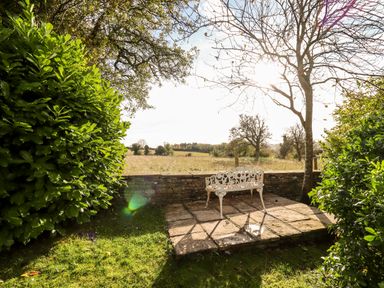 A garden area with a white metal bench on a stone patio surrounded by bushes and trees at The Small House Near Bourton-On-The-Water