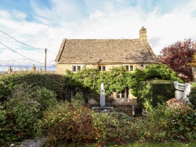 A stone house with a tiled roof covered in greenery and surrounded by various bushes and trees at The Small House Near Bourton-On-The-Water
