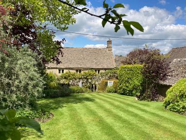 A garden with a mowed lawn and trees with a stone house in the background at The Small House Near Bourton-On-The-Water