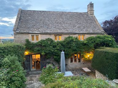 Stone cottage with lit windows and wooden door surrounded by greenery and garden furniture at The Small House Near Bourton-On-The-Water