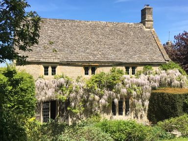 A stone house with climbing plants and flowers on the front surrounded by greenery at The Small House Near Bourton-On-The-Water