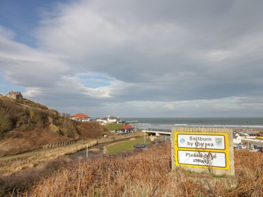 A coastal scene with houses on a hillside a road leading to a bridge and a sea in the background with a sign saying Saltburn by the sea