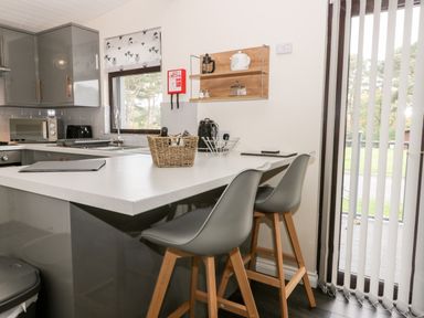A kitchen with grey cabinets a white countertop two bar stools and a window with vertical blinds at Lavender Lodge in Saltburn-By-The-Sea