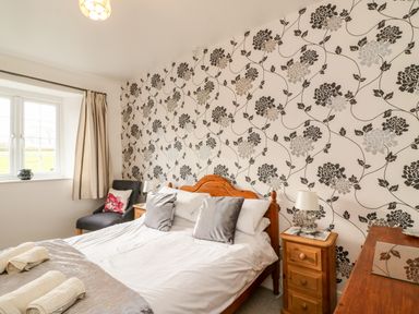 A bedroom with a wooden bed with pillows a bedside table with a lamp a chair by the window and floral patterned wallpaper at Littleham Court Cottage Littleham near Bideford
