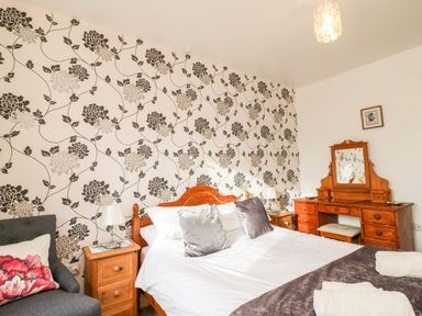 A bedroom with a double bed, bedside tables, a dressing table with mirror, an armchair with a cushion and floral wallpaper at Littleham Court Cottage in Littleham near Bideford