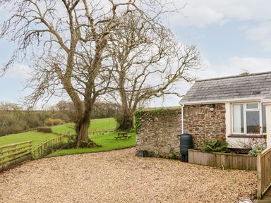 A gravel driveway next to a stone wall and a small building with a window and a rain barrel at Littleham Court Cottage in Littleham near Bideford