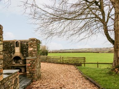 An outdoor area with a stone barbecue and a gravel path next to a fence and a grassy field at Littleham Court Cottage in Littleham near Bideford