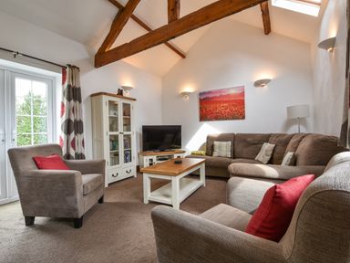 A living room with a brown sectional sofa two armchairs a wooden coffee table a TV on a stand and a cabinet with glass doors at Littleham Court Cottage in Littleham near Bideford