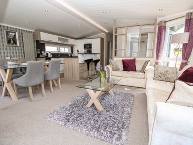 An open plan living room with beige sofas a glass coffee table on a rug dining table with chairs and a kitchen with stools at Crosswinds 67 in Bembridge
