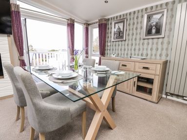 A dining room with a glass table set for four chairs and a wooden sideboard with framed pictures on the wall at Crosswinds 67 in Bembridge
