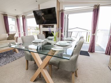 A dining area with a glass table set with plates bowls and glasses next to a living room with a sofa and television at Crosswinds 67 in Bembridge