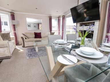 A living room with beige sofas a glass dining table with place settings and a wall-mounted tv at Crosswinds 67 in Bembridge