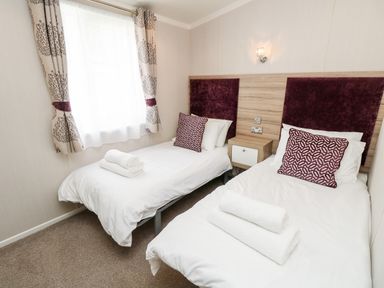 A bedroom with two single beds with white linens and rolled towels by a window with patterned curtains at Crosswinds 67 in Bembridge
