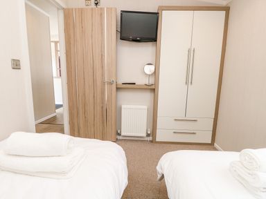 A bedroom with two single beds with white bedding and towels a wooden door a wall-mounted television and a wardrobe at Crosswinds 67 in Bembridge