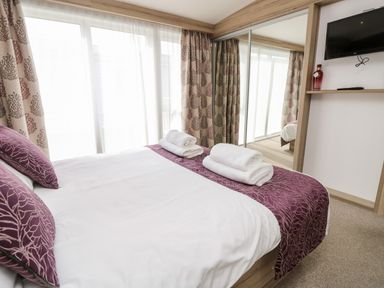 A bedroom with a bed with white sheets and purple pillows and runner towels on the bed curtains and a mirrored closet door a wall mounted TV and a shelf with a red bottle and remote control at Crosswinds 67 in Bembridge