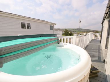 An outdoor hot tub with partially opened cover on a deck beside a white railing overlooking distant hills at Crosswinds 67 in Bembridge