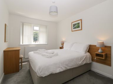 A bedroom with a double bed, bedside lamps, a wooden cabinet with a chair, and a window with vertical blinds at Harcombe House Bungalow 3 in Chudleigh