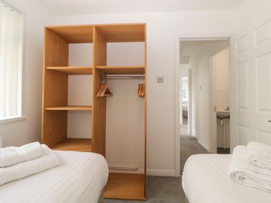 A bedroom with two single beds with white linens and towels a wooden open wardrobe with hangers and a view of a hallway and a bathroom at Harcombe House Bungalow 3 in Chudleigh