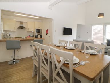 A dining table with six chairs set with plates and glasses next to a kitchen with two bar stools and appliances at Harcombe House Bungalow 4 in Chudleigh