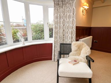 A corner with a white cushioned chair with a fur throw and a book a bay window with a rabbit sculpture and beige curtains at Dant Y Llew in Bow Street Wales