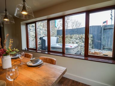 A dining area with a wooden table set with plates bowls and glasses next to a window showing a hot tub and stone wall outside at Dant Y Llew in Bow Street Wales