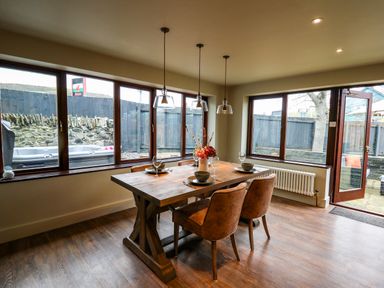 A dining area with a wooden table and four chairs by large windows and a glass door at Dant Y Llew in Bow Street Wales